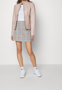 Giacca di pelle rosa chiaro con collo alto, indossata sopra una T-shirt bianca e una gonna a quadri con tonalità di blu, rosa e nero. L'outfit è completato da sneakers bianche.