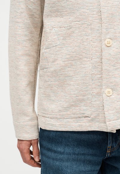 Μπεζ πουκάμισο με μακριά μανίκια και μικρό πολύχρωμο πουά μοτίβο, δύο εμπρόσθιες τσέπες, λευκά κουμπιά, συνδυασμένο με σκούρο μπλε τζιν.