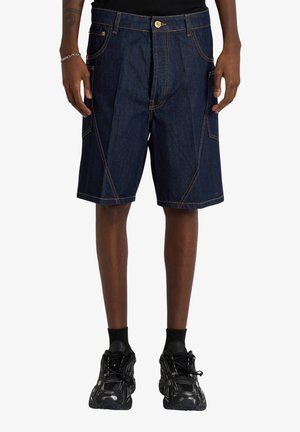 Persona che indossa pantaloncini di denim blu scuro con cuciture arancioni, calzini neri e scarpe sportive nere, mostrata dalla metà della coscia fino ai piedi.