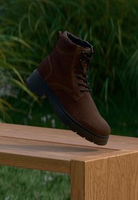 Stivale alla caviglia in suede marrone con suola in gomma nera, design con lacci, punta arrotondata e dettagli di cucitura, posizionato sopra una superficie di legno.