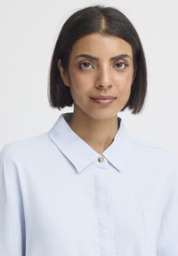 Femme aux cheveux noirs raides portant une chemise bleu clair boutonnée, regardant droit devant avec une expression neutre sur un fond uni.