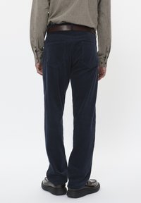 Pantaloni in corduroy blu navy con taglio dritto, tasche posteriori e tessuto testurizzato. Abbinati a una camicia beige e a una cintura in pelle marrone.
