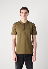 Olive zaļas polo kreklis ar īsām piedurknēm, ar rāvējslēdzēja apkakli un smalku logotipa detaļu kreisajā krūtī. Izgatavots no gludas, elpojošas auduma.