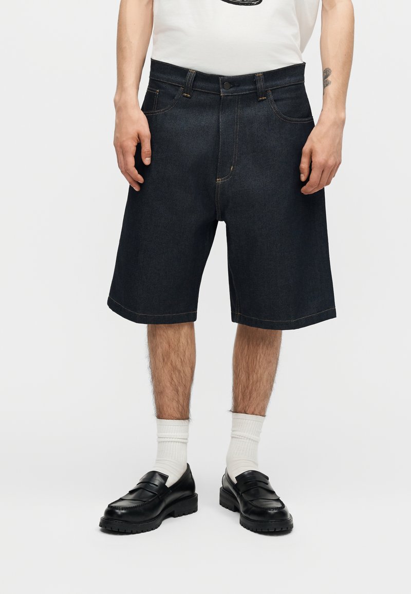 Uomo che indossa pantaloncini di jeans scuri fino al ginocchio, calzini bianchi, mocassini neri e una maglietta bianca con un tatuaggio visibile sul braccio.