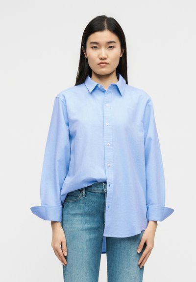 Γυναίκα που φοράει ανοιχτό μπλε πουκάμισο με κουμπιά και μανίκια που έχουν γυριστεί, καθώς και μπλε τζιν, στέκεται μπροστά σε ένα απλό φωτεινό φόντο.