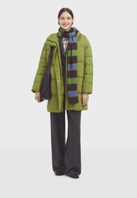 Grøn puffjakke, stribet halstørklæde i blå, grøn og brun, grå ternet skjorte under, sort taske, brede grå bukser, glat tekstur.