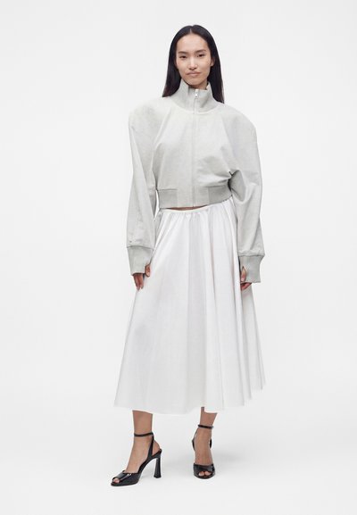 Femme aux longs cheveux foncés portant un pull gris clair zippé, une jupe midi blanche et des sandales noires à talons hauts, debout devant un fond blanc uni.