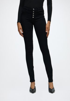 Slim fit sorte bukser med fem synlige knapper foran, båret med sort langærmet top og sorte spidse sko på ensfarvet baggrund.
