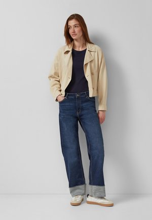 Ženska stoji z rokami v žepih, oblečena v bež jakno, mornarsko modro majico, zavihane modre kavbojke in bele superge na enostavni svetlo sivi podlagi.