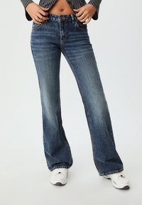 Flared blue denim jeans with a faded wash, stitched detailing, and a high waist. Worn with white athletic shoes.