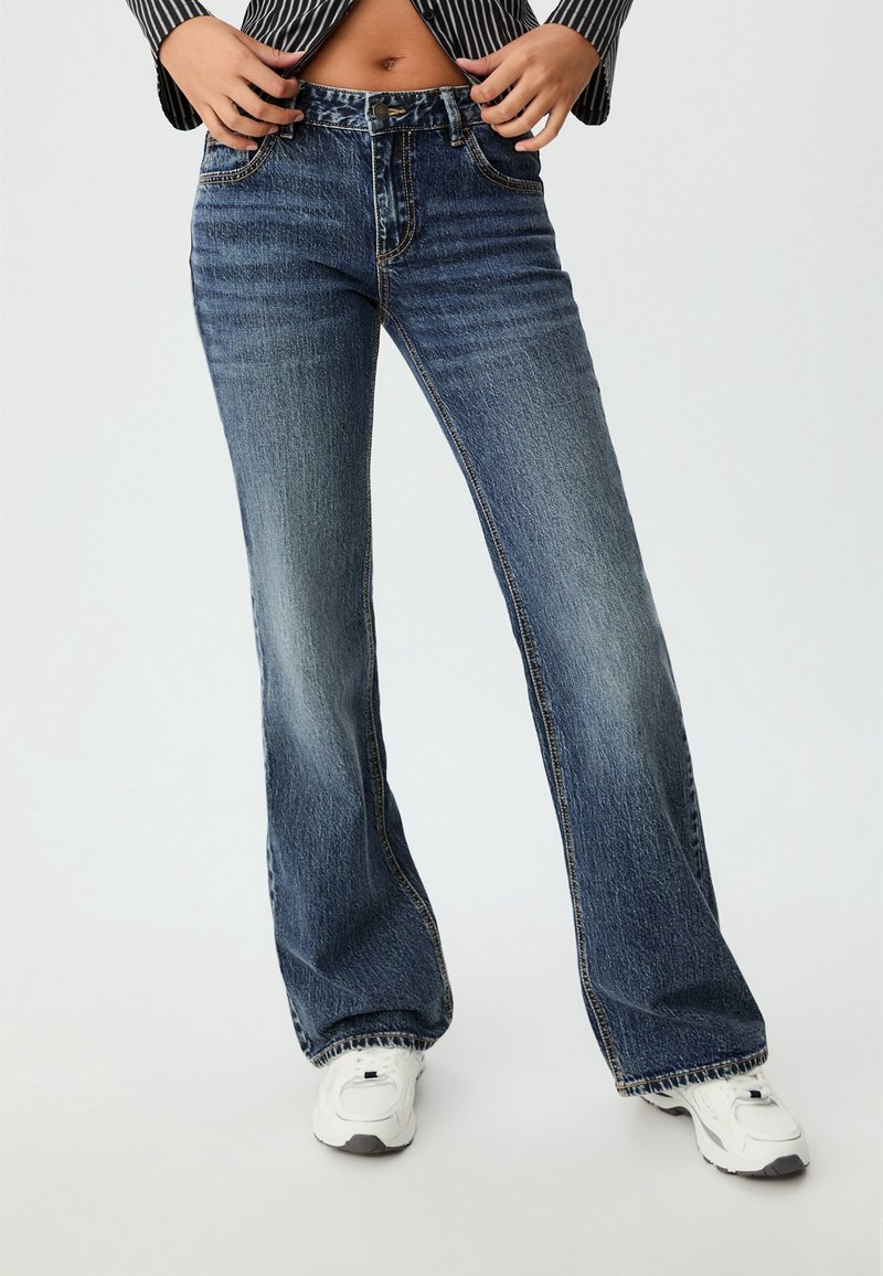 Flared blue denim jeans with a faded wash, stitched detailing, and a high waist. Worn with white athletic shoes.