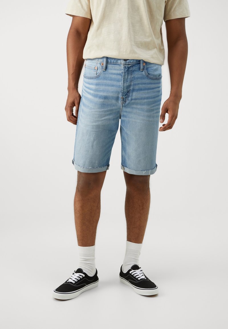 Homme portant un short en jean bleu clair, un t-shirt beige, des chaussettes blanches et des baskets noires basses sur un fond blanc uni.