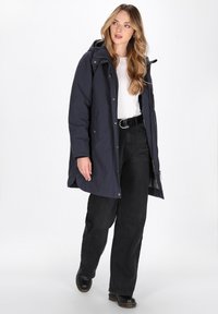 Jonge vrouw met lang golvend haar, gekleed in een marineblauwe capuchonjas, witte top, zwarte riem, wijde zwarte broek en zwarte laarzen, staande voor een witte achtergrond.