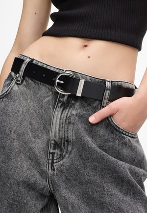Μαύρη δερμάτινη ζώνη με ασημένια αγκράφα, φορεμένη σε ψηλόμεσα, ξεθωριασμένα μαύρα τζιν παντελόνια με χαλαρή εφαρμογή και αισθητή υφή.