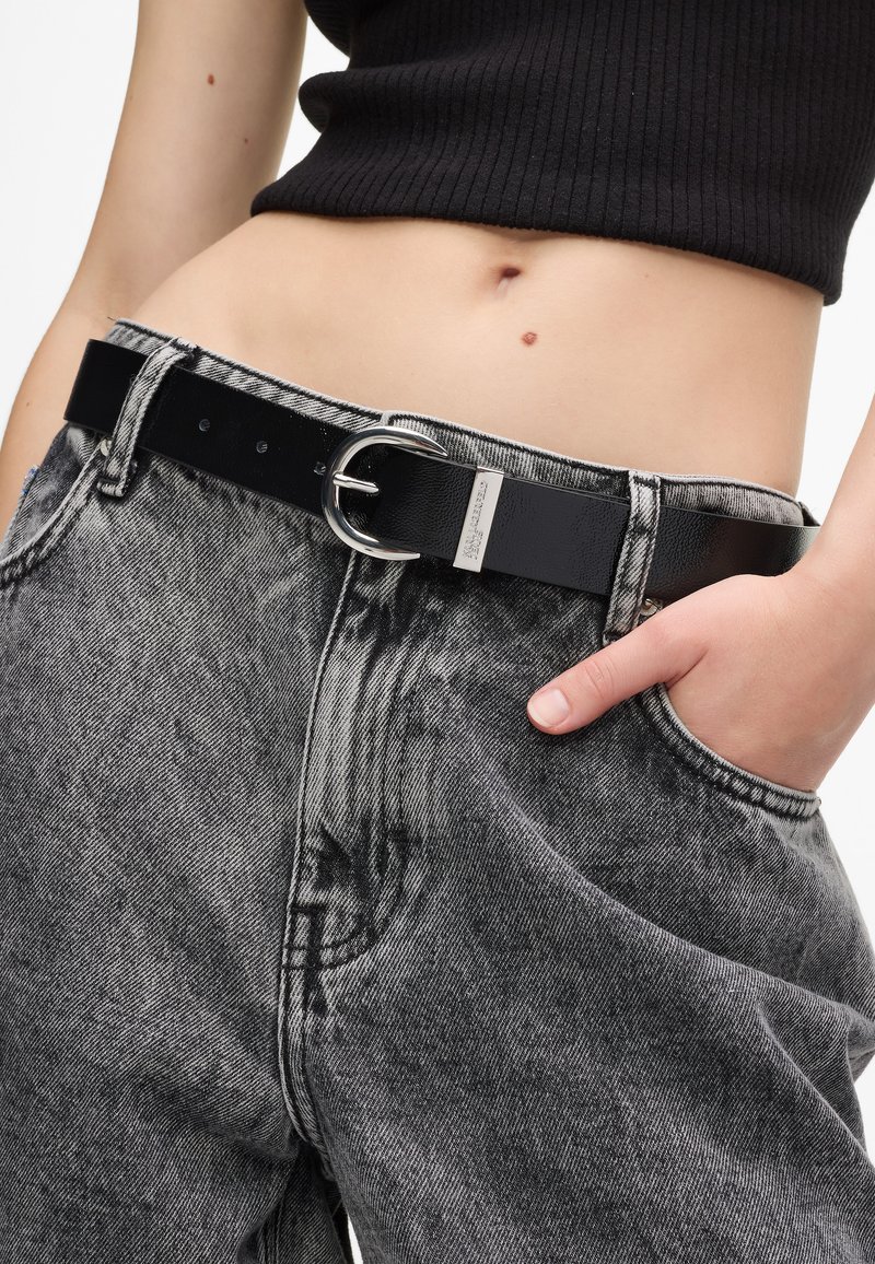 Juodas odinis diržas su sidabriniu sagtimi, dėvimas ant aukšto liemens, išblukusių juodų džinsų su laisvu siluetu ir prašmatnia tekstūra.