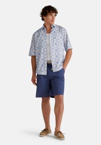Camisa de manga corta, de color azul claro con patrones circulares blancos, combinada con pantalones cortos azul marino. Unos zapatos de barco beige completan el look.