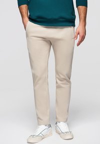 Hombre con pantalones ajustados beige, zapatillas blancas con detalles negros y sudadera verde oscuro, de pie con las manos en los bolsillos.