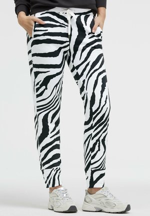 Person trägt schwarz-weiß gestreifte Jogginghosen im Zebramuster mit Kordelzugbund und weiße Sportschuhe, Hände in den Taschen.