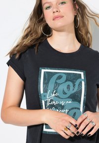 Mujer con cabello ondulado, delineador verde, pendientes de aro y esmalte de uñas negro, lleva una camiseta negra con un gráfico azul y anillos plateados en los dedos.