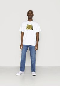 Biała bawełniana koszulka z graficznym wzorem zawierającym żółte i czarne elementy. W połączeniu z jasnoniebieskimi dżinsami i białymi trampkami.