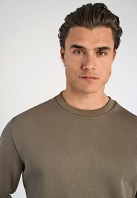 Camisa verde oliva de manga larga con cuello redondo, hecha de una tela suave y texturizada con un diseño ajustado. Simple y sin adornos.