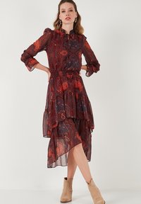 Punainen ja sininen paisley-kuvioinen mekko, joka on tehty läpinäkyvästä kankaasta, rypytetyllä helmalla, kerätyllä vyötäröllä ja pitkillä hihoilla. Tyylitelty vaaleanruskeilla nilkkureilla.