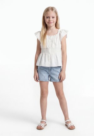 Νεαρό κορίτσι με μακριά ξανθά μαλλιά που φοράει λευκό αμάνικο μπλουζάκι, ανοιχτό μπλε τζιν σορτς και λευκά σανδάλια, στέκεται μπροστά σε λευκό φόντο.