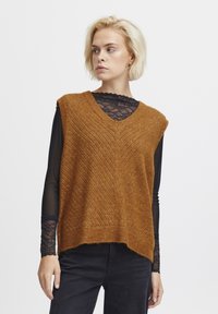 Ärmelloser V-Ausschnitt-Pullover in warmem Braun, mit einem Zickzack-Strickmuster und einem wellenförmigen Saum, über einem schwarzen Spitzentop getragen.
