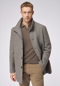 Grijze wollen jas met een gestructureerde afwerking, staande kraag en bruin knopen, gedragen over een bruine polo en gecombineerd met beige broek.