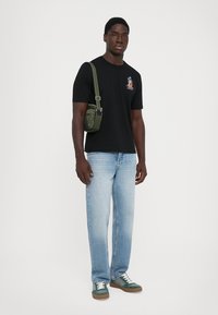 Man wearing black graphic t-shirt, light blue jeans, green crossbody bag, and green sneakers with white laces, standing against white background.