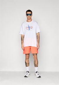 Mann med reflekterende sports solbriller, hvit grafisk t-skjorte, lyse orange shorts, hvite sokker og hvite løpet sko, som står mot en enkel vegg.