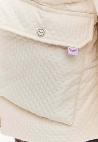 Béžová prošívaná bunda s prošívanou texturou, klopovou kapsou na patentní knoflík a malou štítkem s červenými grafickými akcenty.