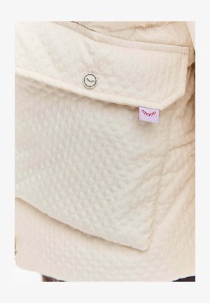 Beżowa pikowana kurtka z wyściełanym materiałem, kieszenią na patkę z zatrzaskiem i małą metką z czerwonymi graficznymi akcentami.