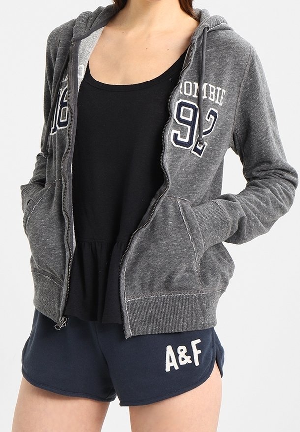 Sudadera gris con cremallera y bolsillo frontal, que presenta el texto "ABERCROMBIE 92" en blanco. Lleva puesta sobre un top negro sin mangas y pantalones cortos azul marino con bordado "A&F".