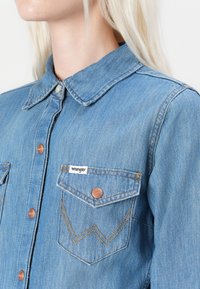 Blue denim shirt with a collar, button closure, and two chest pockets featuring a "Wrangler" label and copper-colored hardware.