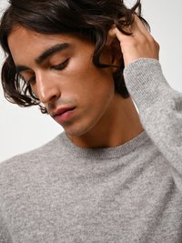 Maglione grigio lavorato a maglia con collo rotondo e polsini a coste, caratterizzato da una texture morbida. Il modello ha capelli scuri e ondulati e un'espressione neutra.