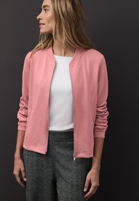 Světlé růžové zipové sako s žebrovaným límcem a manžetami, doplněné bílým topem a texturovanými šedými kalhotami. Měkká látka, ležérní design.