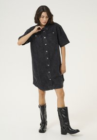 Vestido camisa de mezclilla negra con mangas cortas, cierre de botones en la parte delantera, dos bolsillos en el pecho y un ajuste relajado, combinado con botas altas negras brillantes.