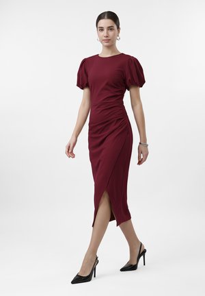 Femme marchant portant une robe midi bordeaux ajustée avec des manches ballon et des chaussures à talons aiguilles noires à bride arrière sur fond blanc uni.