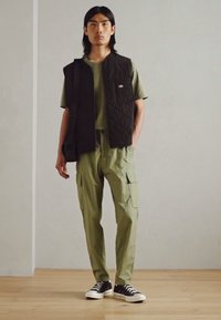 Czarna pikowana kamizelka na zielonym T-shircie, w połączeniu z spodniami cargo. Czarne sneakersy z białymi podeszwami. Prosty design, luźny krój, funkcjonalne kieszenie.