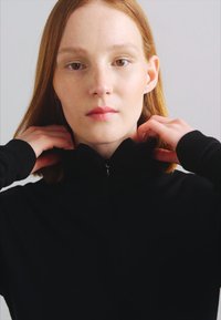 Mustapaita, jossa on korkea kaulus ja vetoketju, valmistettu pehmeästä materiaalista. Malli säätää kaulusta, esitellen tyylikästä muotoilua ja istuvaa leikkausta.