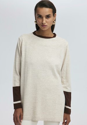 Žena v béžovém svetru s dlouhými rukávy, tmavě hnědými manžetami a výstřihem, stojící před jednoduchým světle šedým pozadím.