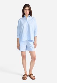 Chemise en coton bleu clair à boutons avec des manches longues, associée à un short bleu clair assorti. Portée avec des sandales marron. Design simple.