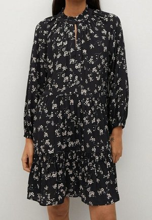 Woman wearing a black long-sleeve dress with white floral print, gathered waist, high neckline, and a small front keyhole opening.