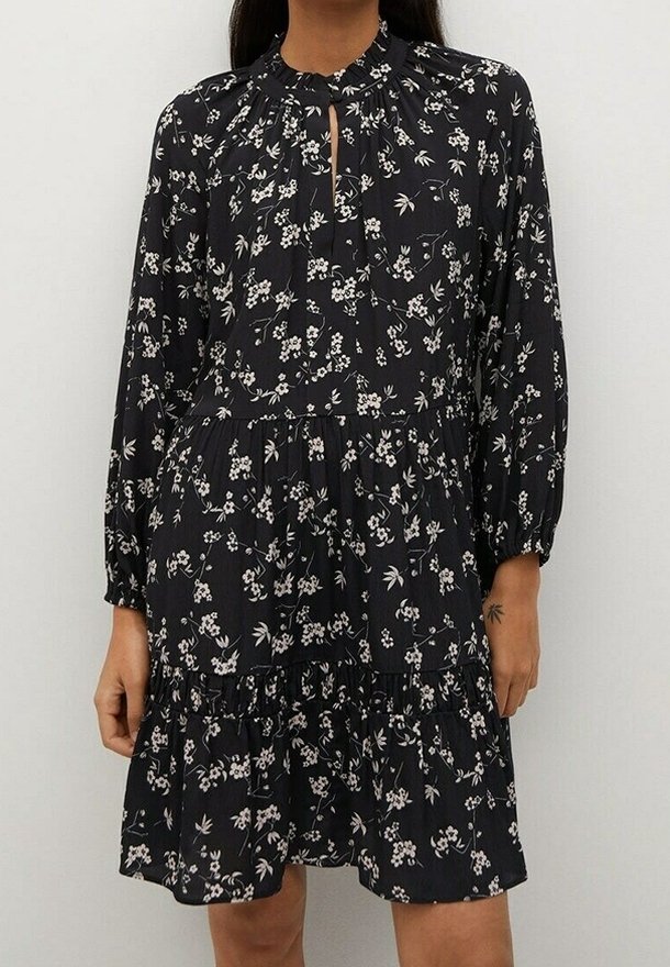 Femme portant une robe noire à manches longues avec imprimé floral blanc, taille froncée, col haut et petite ouverture en forme de goutte à l'avant.