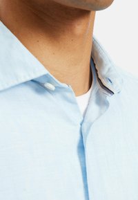 Camicia azzurro chiaro con bottoni, realizzata in un tessuto texturizzato, dotata di un colletto con motivo interno a contrasto e una leggera lucentezza.