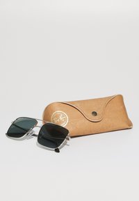Ray-Ban aurinkolasit mustilla tummennetuilla linsseillä, hopeisessa metallikehyksessä ja läpinäkyvillä temppelivihjeillä, asetettuna tanvärisen nahkakotelon viereen, jossa on napsautuskansilukko.
