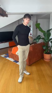 Mladý muž v čiernej dlhorukávovej košeli a béžových nohaviciach stojí s rukami v kapsách v obývacej izbe s hnedou pohovkou a veľkou kvetináčovou rastlinou.