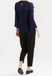 Mujer vistiendo una blusa azul marino con un yugo de encaje y mangas acampanadas, pantalones negros hasta el tobillo, y mocasines negros, vista desde atrás.