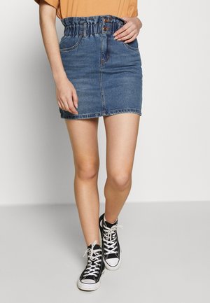 Kobieta ubrana w niebieską spódnicę typu paperbag z denimu, pomarańczowy top i czarne wysokie trampki, chodzi po jasnoszarej podłodze na tle białej ściany.